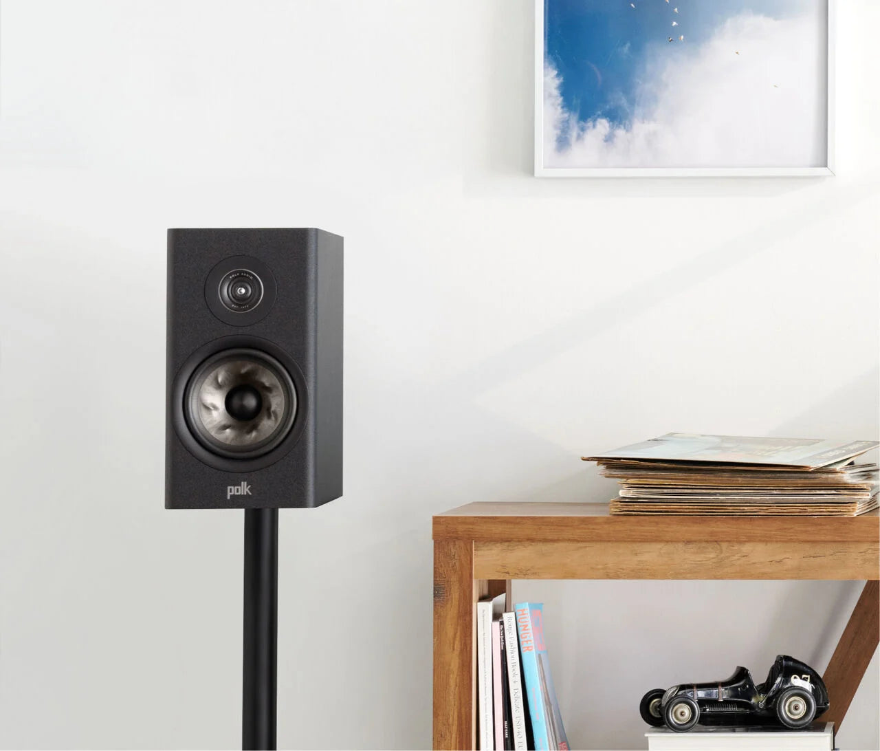 Polk Audio Reserve R100 Boxe de raft negre, vedere frontală, montate pe suport lângă masă din lemn.