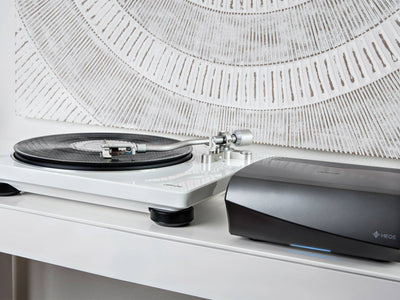 Pick-up Denon DP-400 alb, vedere laterală, cu platan vinil și braț metalic vizibil. Alături este amplificatorul Denon HEOS.