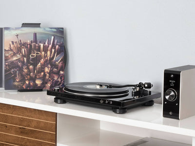 Pick-up Denon DP-450USB negru, vedere laterală, cu braț și platan vizibile, amplasat pe raft alb. Dispozitiv audio în fundal.