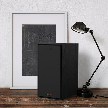 Boxă activă Klipsch R-50PM neagră, vedere frontală cu grilaj montat, amplasată lângă lampă și tablou.